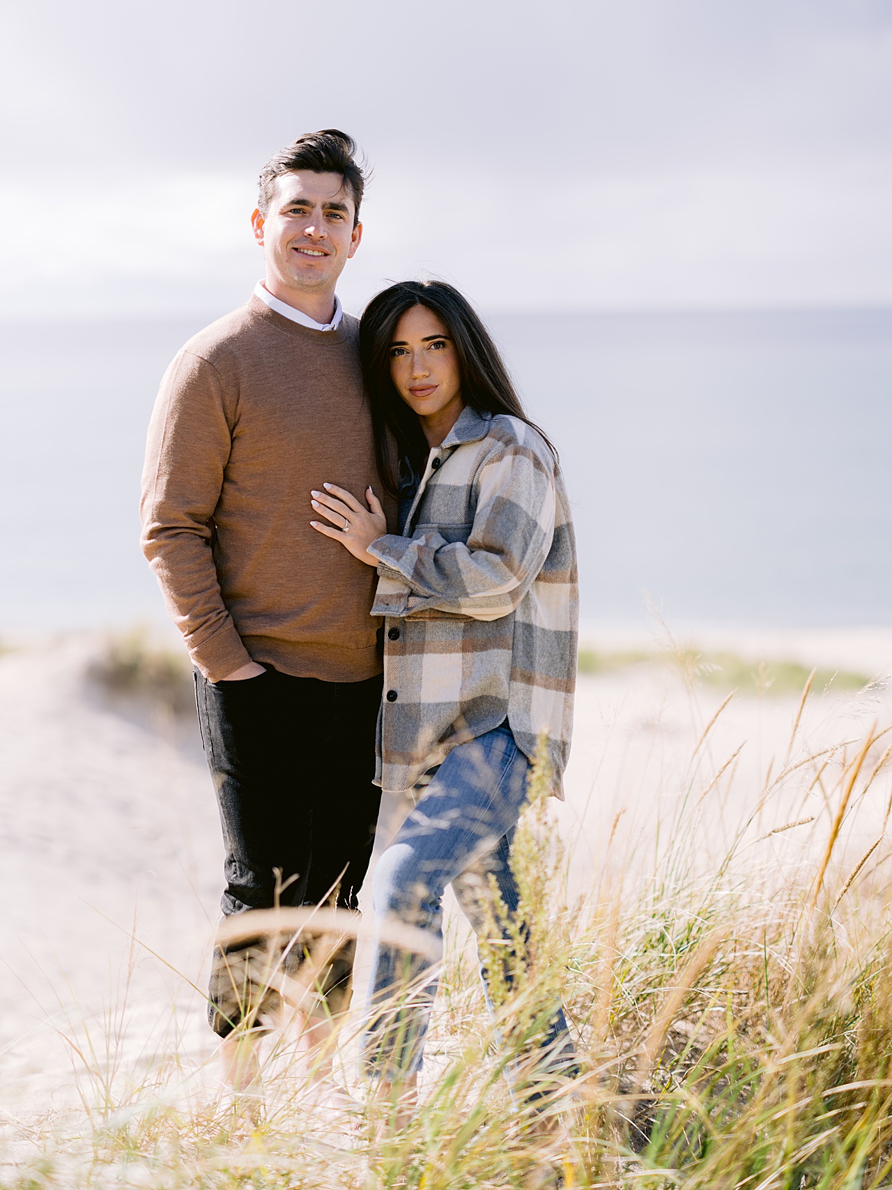 An engaged couple poses together next to tall grass on a sandy beach in Petoskey
