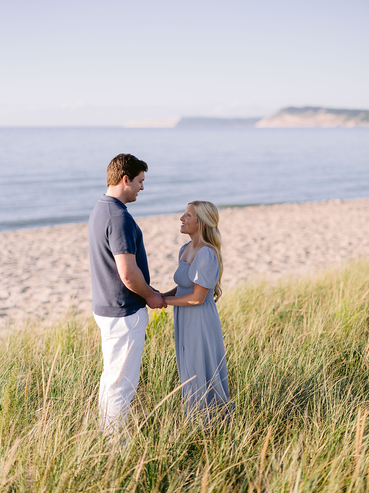 An engaged couple holding hands and looking at each other in dune grass with Lake Michigan in the background on a sunny day