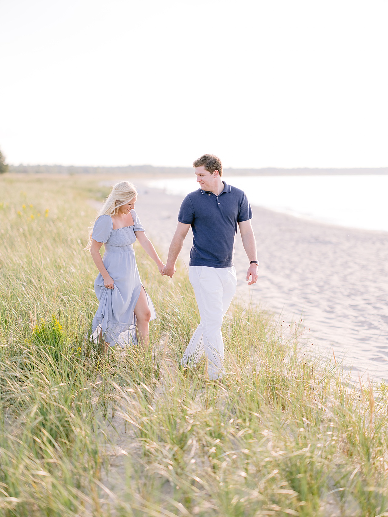 An engaged couple walking through dune grass towards Lake Michigan