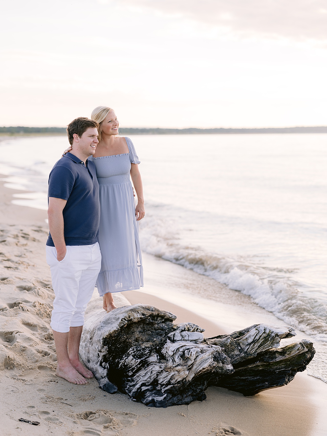 A woman in a long light blue dress standing on a large washed up tree along a lakeshore and leaning onto her fiancé