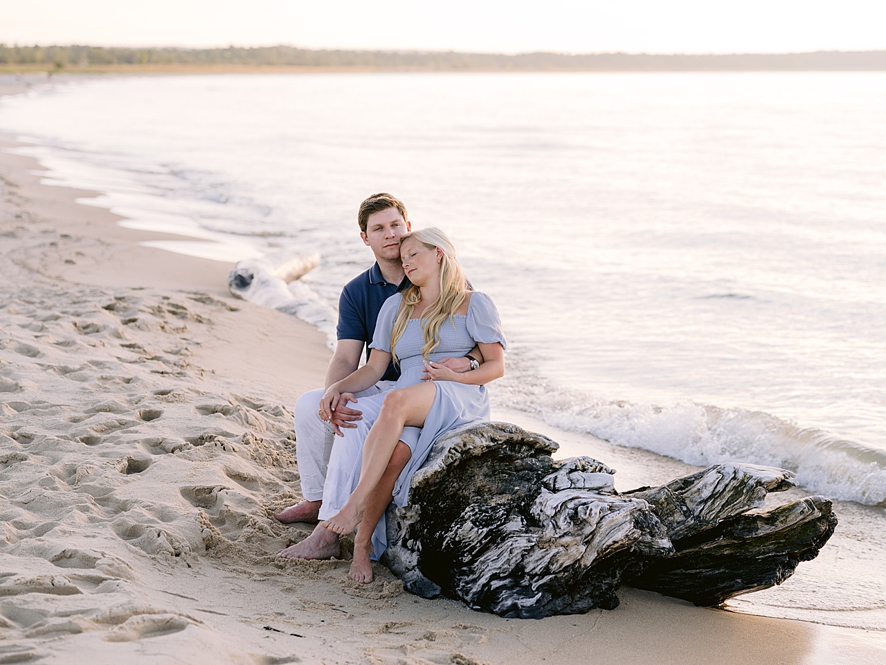 An engaged couple snuggling up together on a large driftwood tree as waves crash onto it