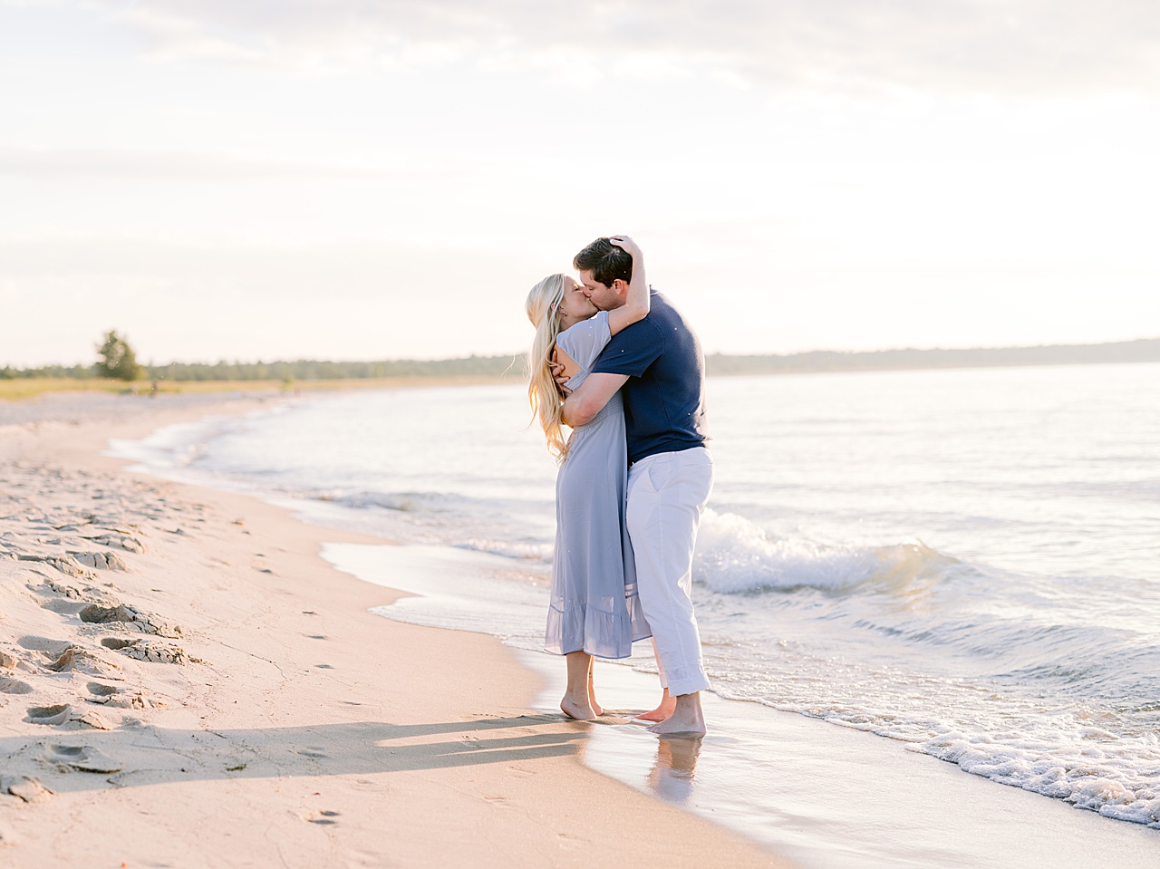 An engaged man and woman holding each other and kissing while their feet are soaking in the freshwater lake in Michigan