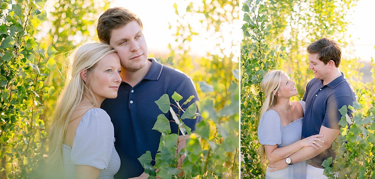 A man and woman fiancé smile at each other while standing in an area surrounded by bright green trees