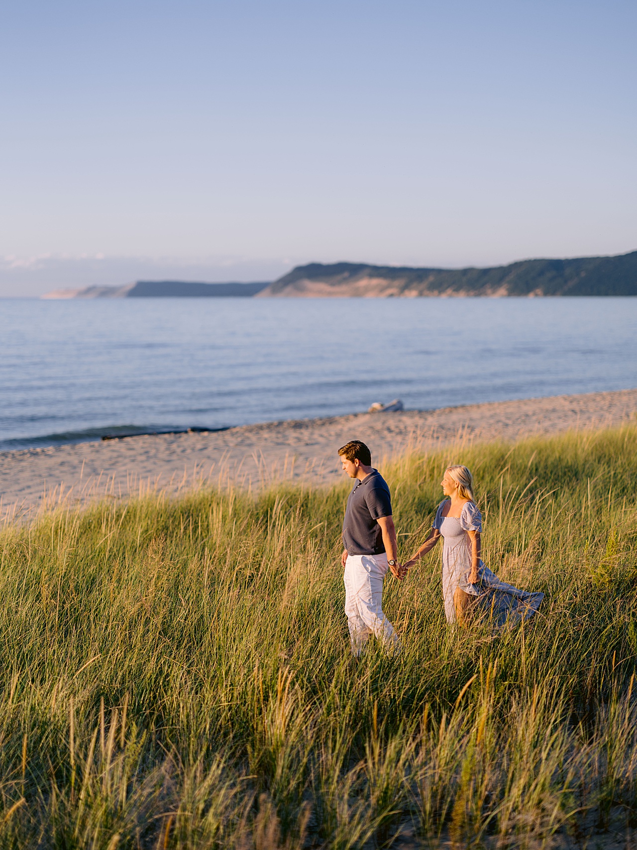 Engaged couple walking hand in hand through dune grass at sunset with Sleeping Bear Dunes in Honor, Michigan