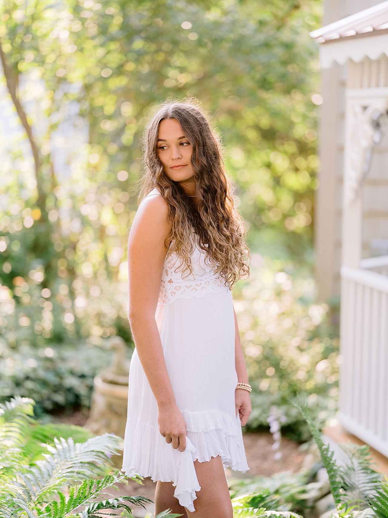 A high school girl stands in a group of ferns while wearing a white dress