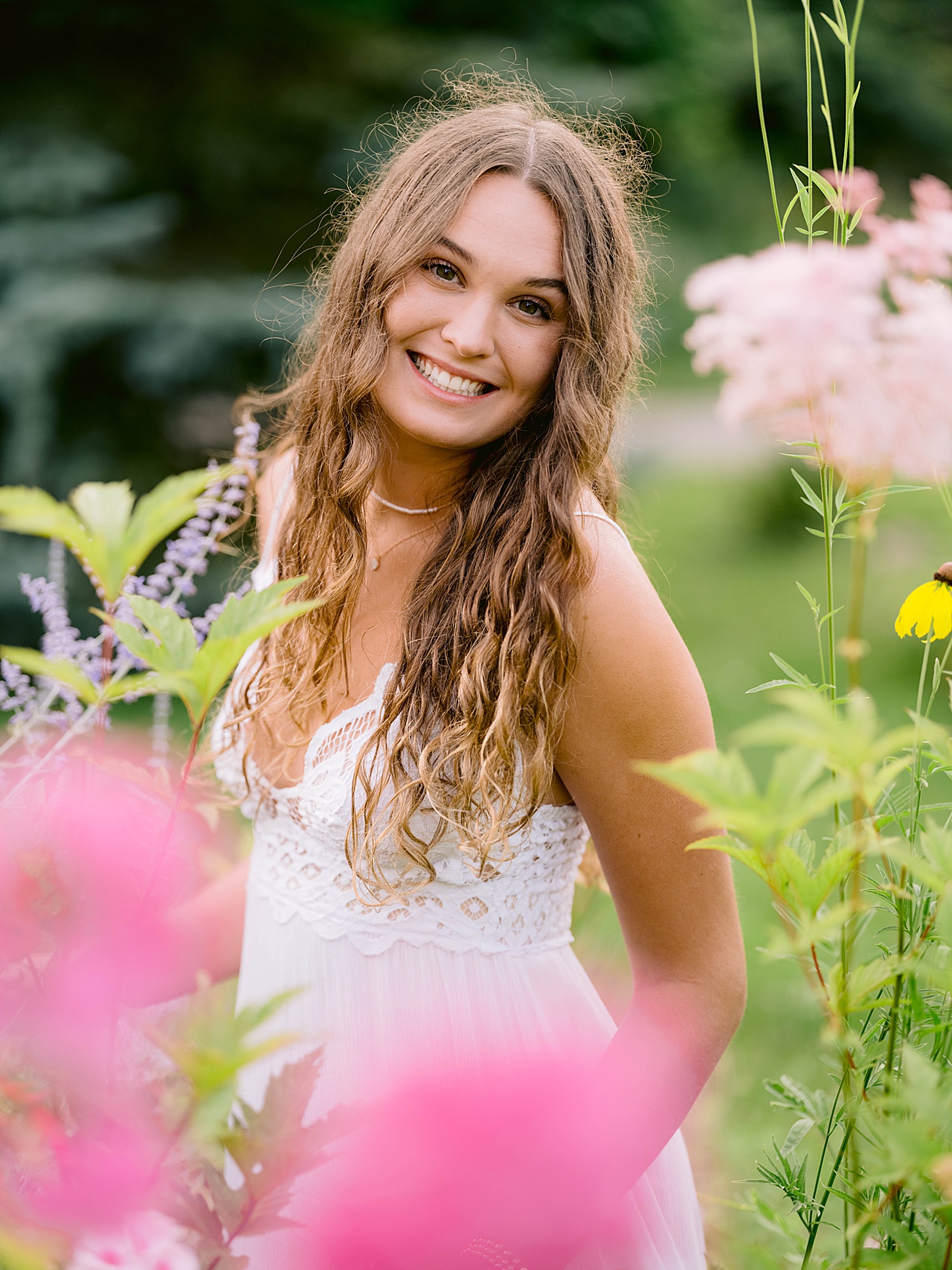 A girl with curly brown hair smiles among tall flowers for senior portraits in michigan