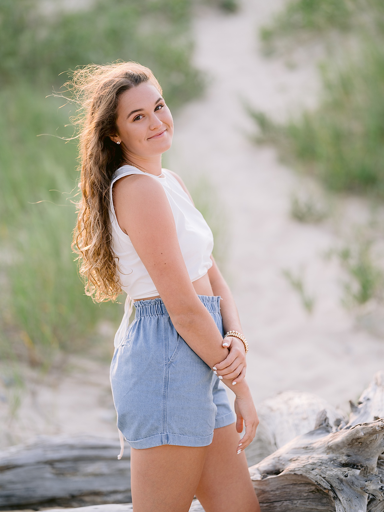 A girl stands on a sandy beach and poses for senior photos beside a large piece of driftwood