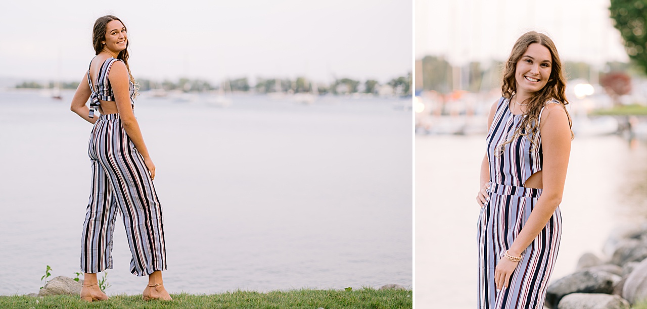 A girl poses beside a lake for harbor springs senior portraits in a striped jumpsuit