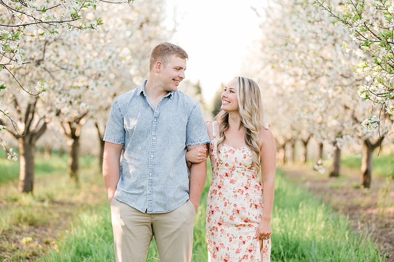 An engaged couple look at each other and smile lovingly in an orchard of cherry blossoms