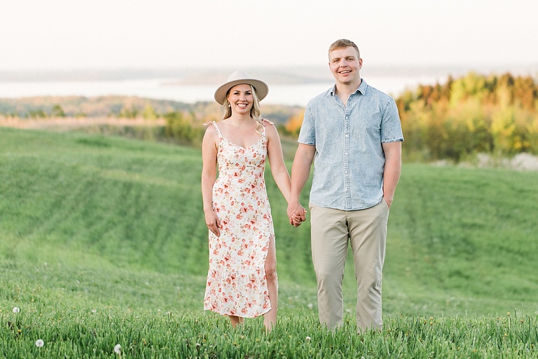 A man and woman hold hands and pose for portraits on a hill overlooking a field, forest and lake