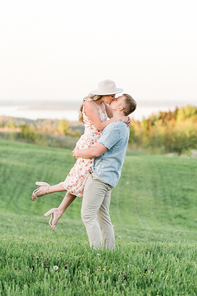 A man lifts a woman off the ground and the two kiss while standing in a grassy field in northern Michigan