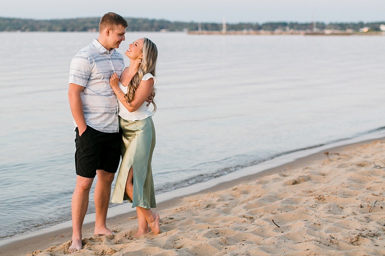 A woman smiles up at her fiancé while they stand barefoot in the sand holding each other