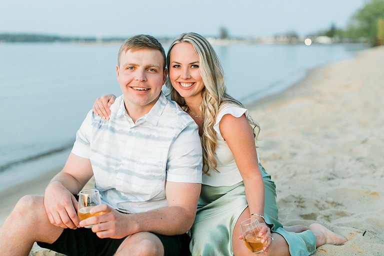 An engaged couple sits on a sandy beach while snuggled up holding glasses of sparkling wine