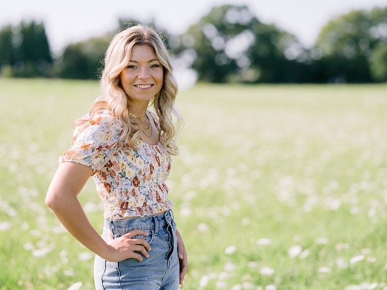 A young woman stands smiling for senior portraits with a hand on her hip