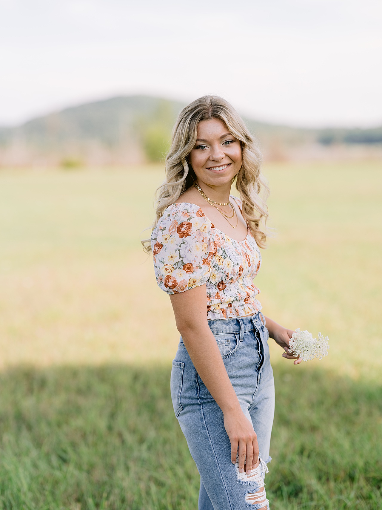 A high school senior stands in a field holding white flowers in her hand during a photo session
