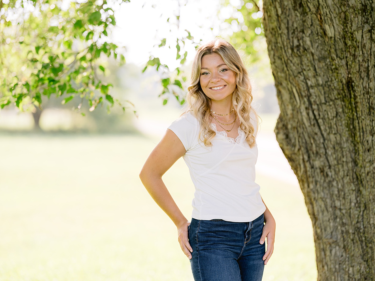A girl stands near a tree trunk under the leaves of a tree branch in michigan