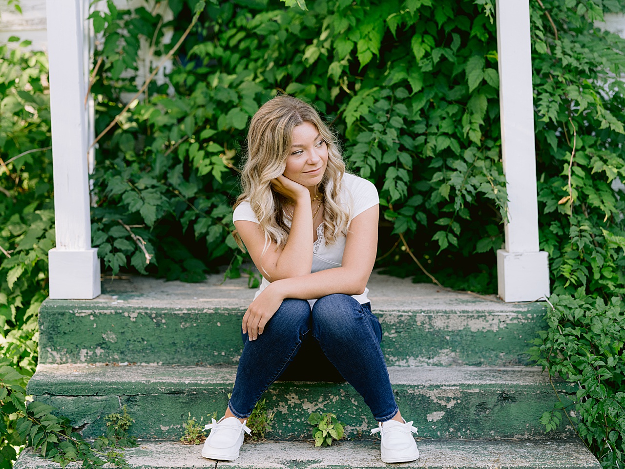 A high school senior sits on the steps of an old building with overgrown foliage behind her