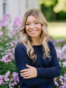 A high school senior girl in a navy blue sweater smiles and poses in front of purple flowers