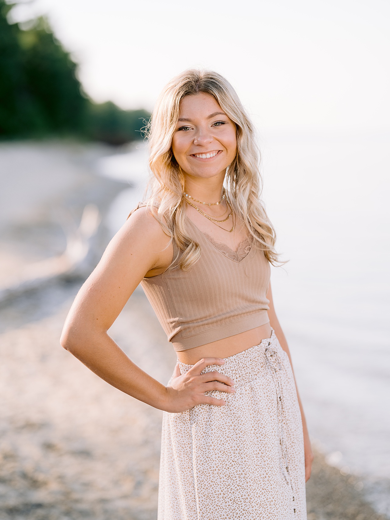 A girl stands with hand on hip and smiles with Lake Michigan shoreline behind her