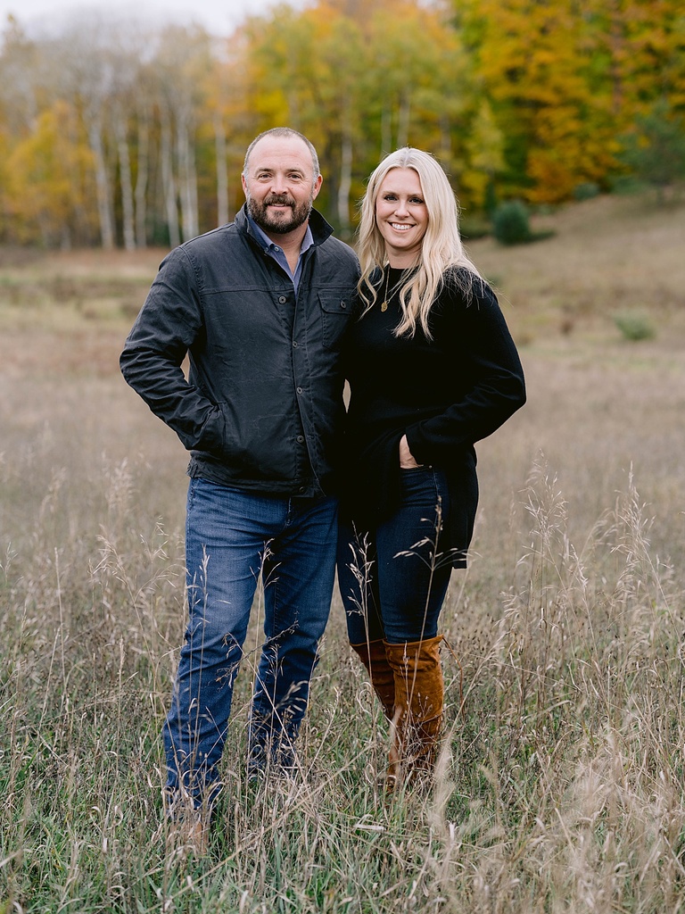 A couple stands next to each other in a field and smiles for michigan fall family portraits