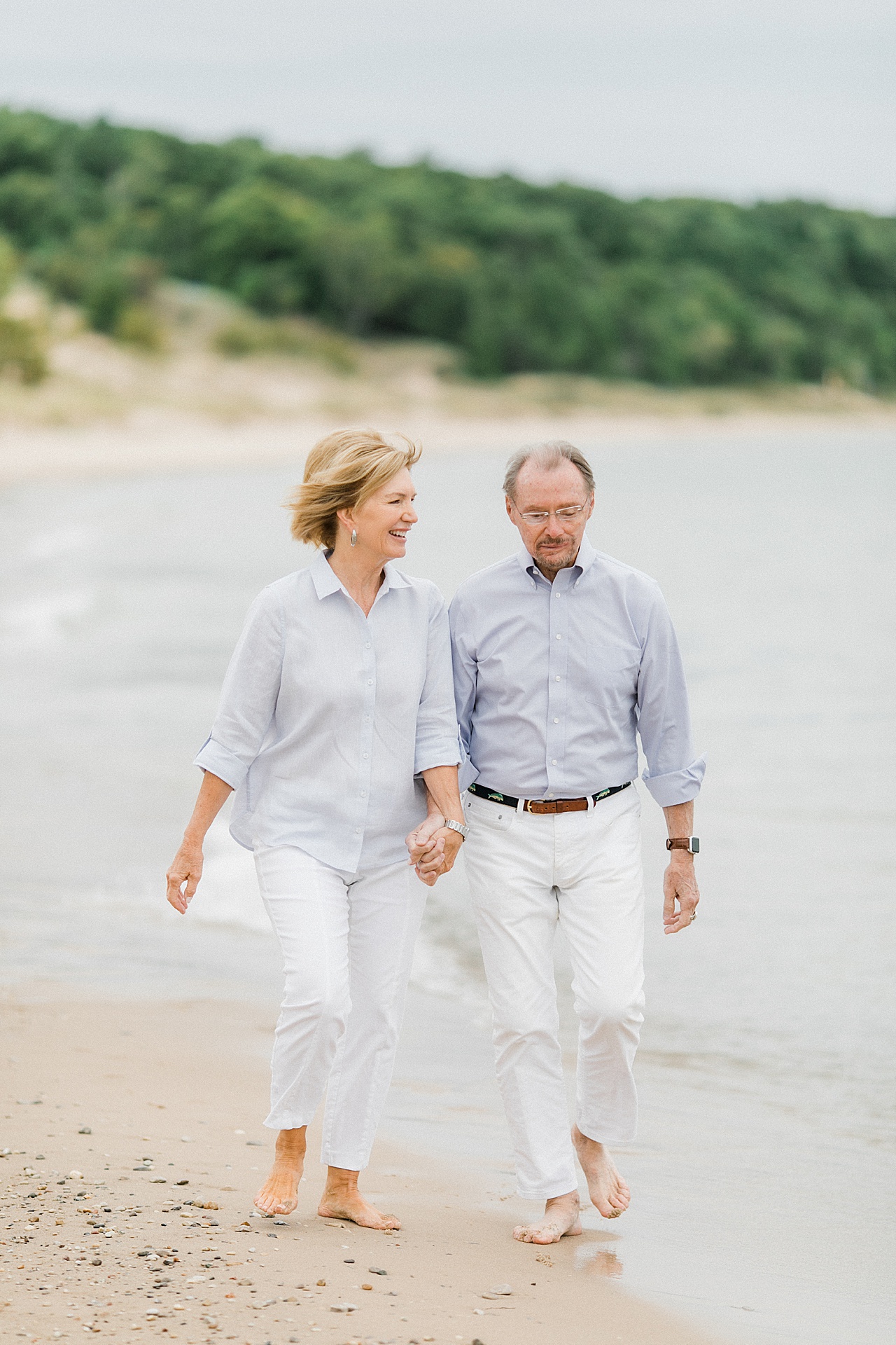 A distinguished older couple walk up a beach together holding hands and smiling