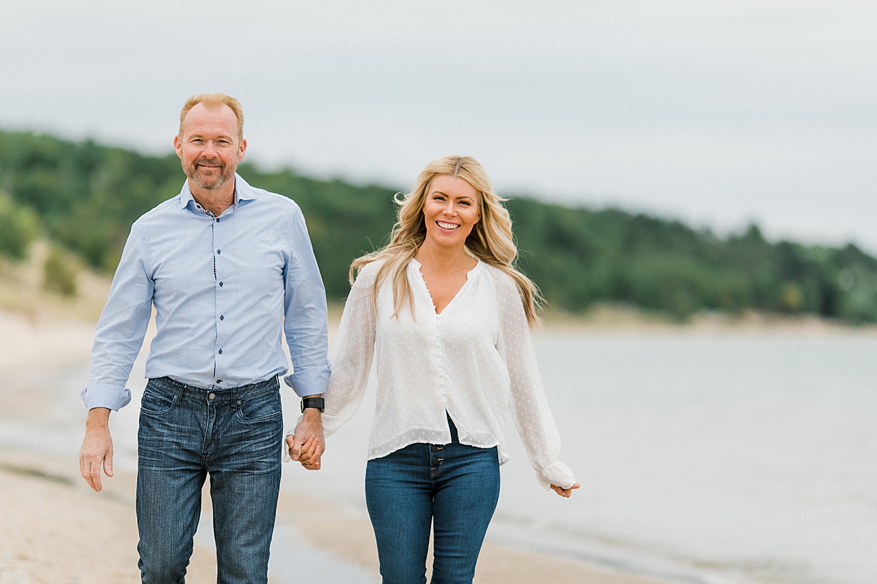 A happy couple walks up a beach holding hands while the wind blows there hair