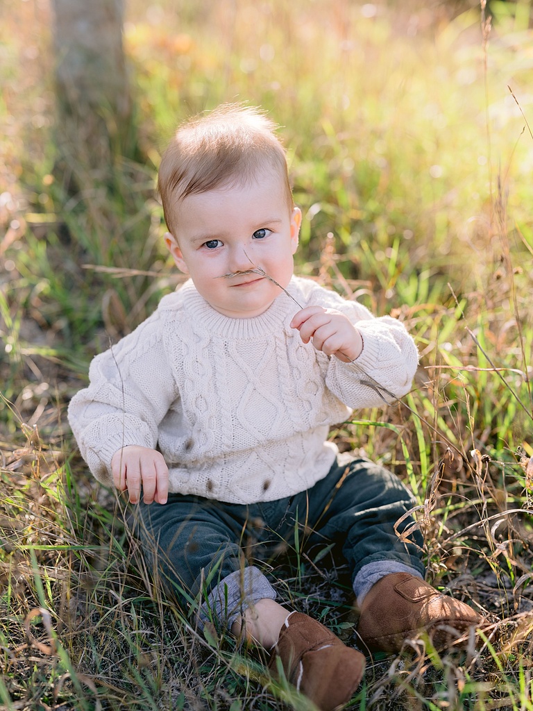 A one year old boy in a knit off white sweater, blue jeans, and brown shoes playing with grass in a field